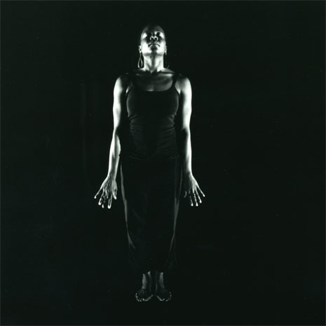 performer aimee meredith cox, standing upright, her arms stretched downwards, in dark theatre