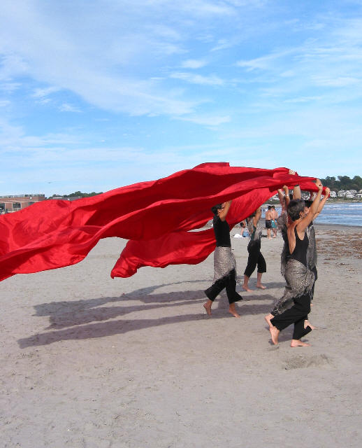 performers moving across a beach, with red clohtes billowing behind
